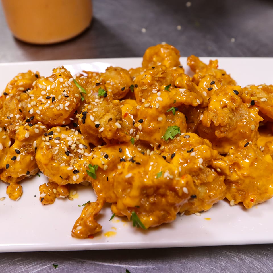 Crispy chicken pieces coated in orange sauce, topped with white and black sesame seeds and cilantro on a white platter.