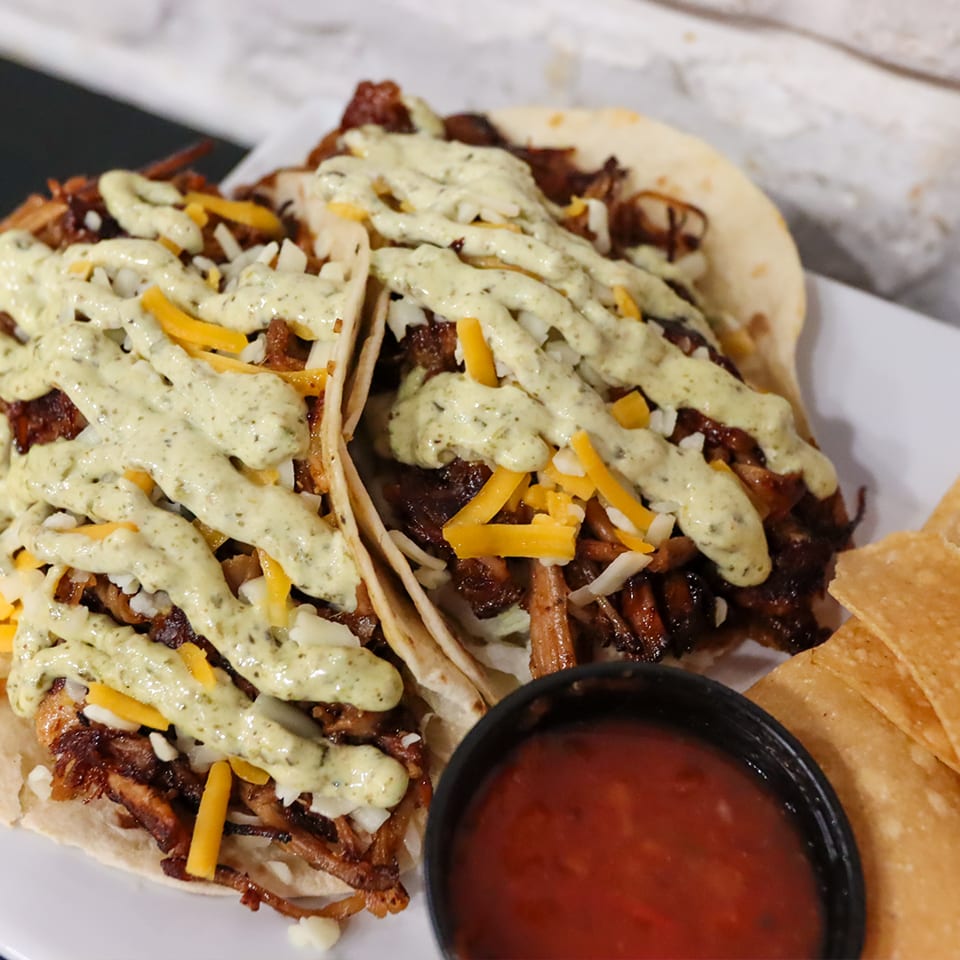 Two tacos filled with meat, cheese, and a creamy green sauce, served with salsa and chips.