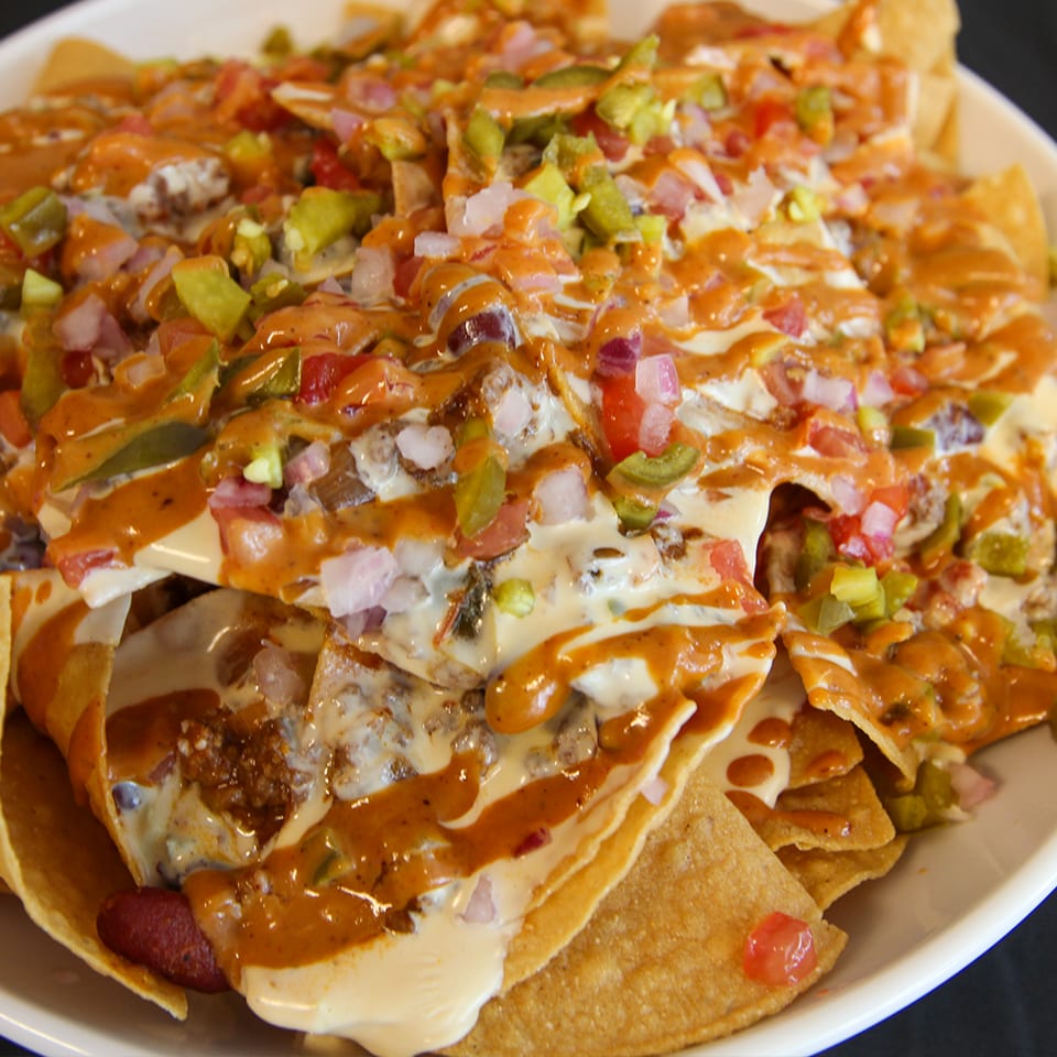 Loaded nachos topped with melted cheese, seasoned meat, diced tomatoes, onions, peppers, and creamy orange sauce on a plate.