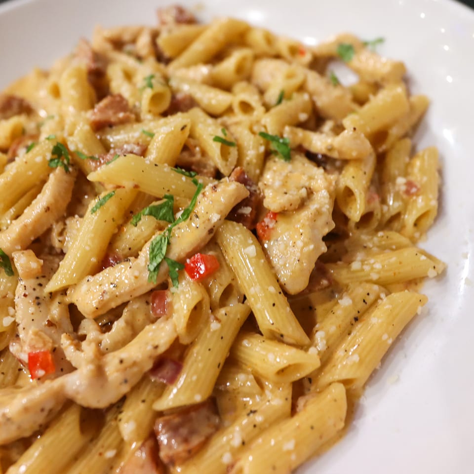 Creamy penne pasta with chicken, bacon, red peppers, herbs, and cracked black pepper on a white plate.