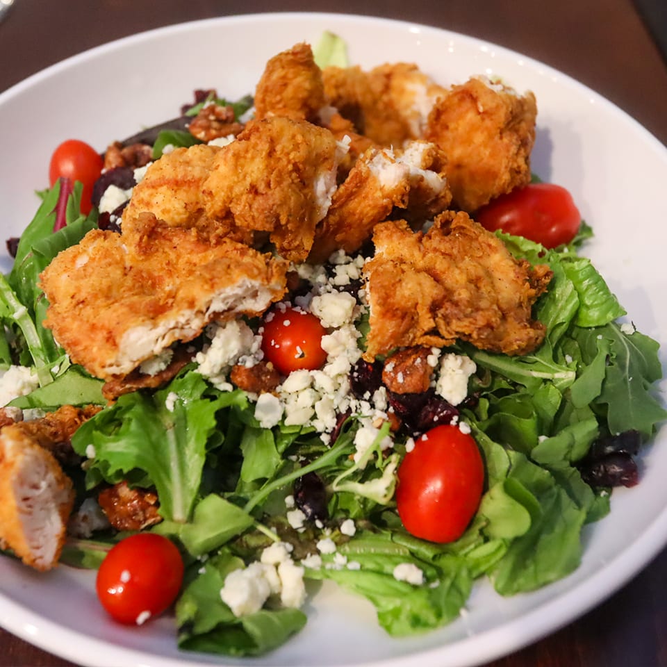 Crispy fried chicken pieces atop mixed greens with cherry tomatoes, blue cheese crumbles, and nuts on a white plate.