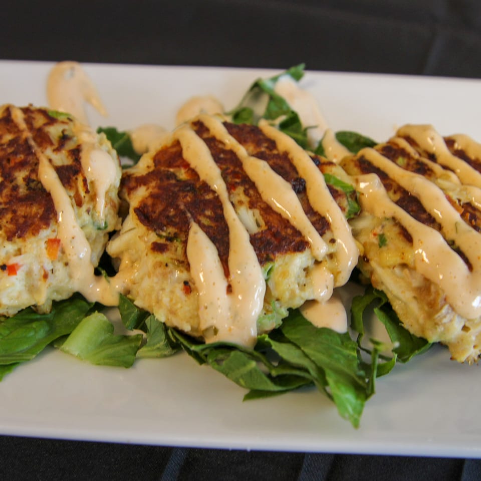 Three golden-brown crab cakes drizzled with creamy sauce, served on a bed of fresh green lettuce.